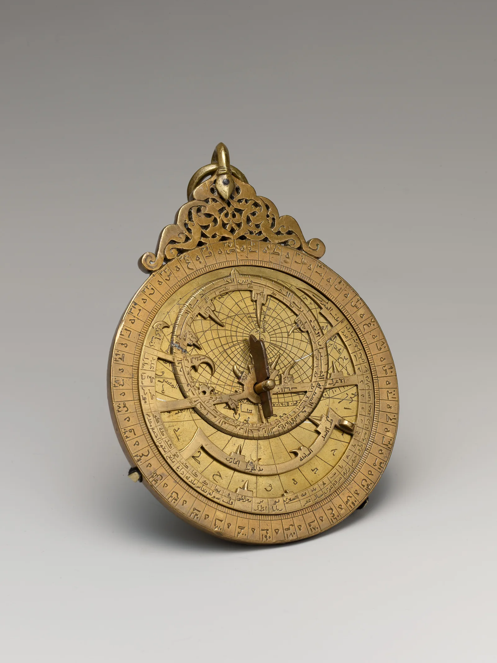 Round brass astrolabe with suspension ring, pierced rete, engraved scales, Arabic inscriptions, and movable pointer.