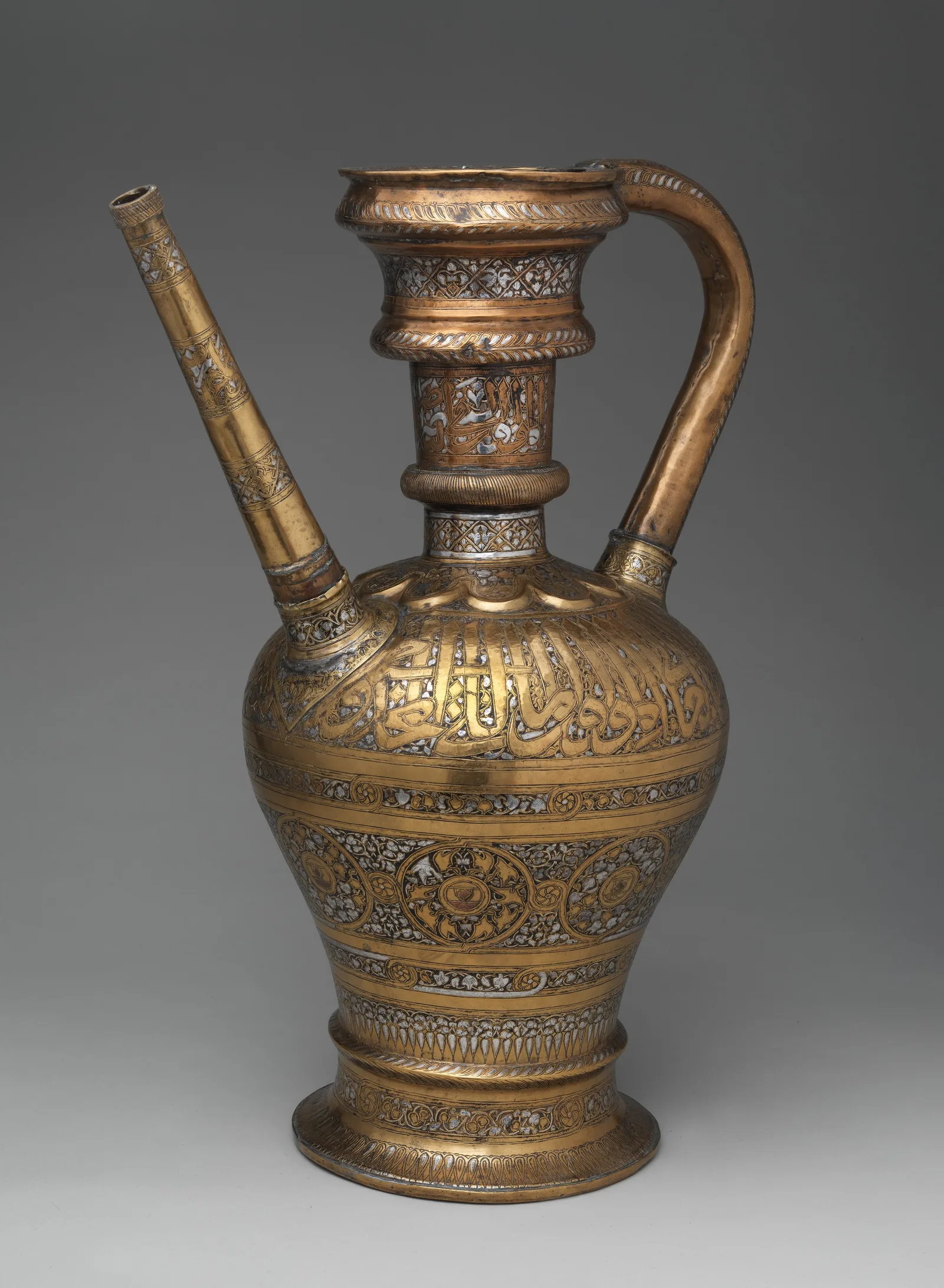 Tall brass ewer with long spout, rounded handle, engraved bands, silver inlay, copper accents, and dark compound in recessed ornament.